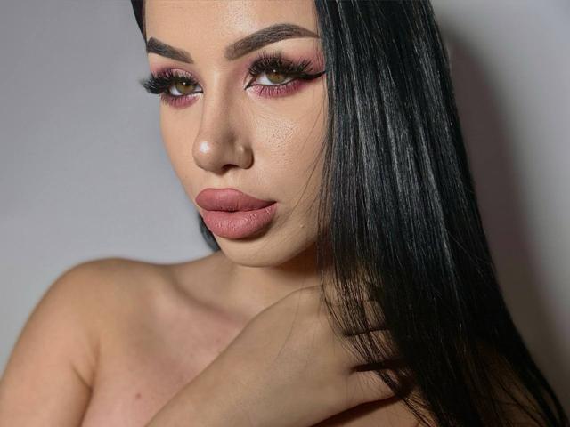 JennaDay has long black hair, pink eyeshadow, thick eyelashes, and pouty pink lips. Her hand touches her neck, showing off her bare shoulders. She has a sexy, sultry look.