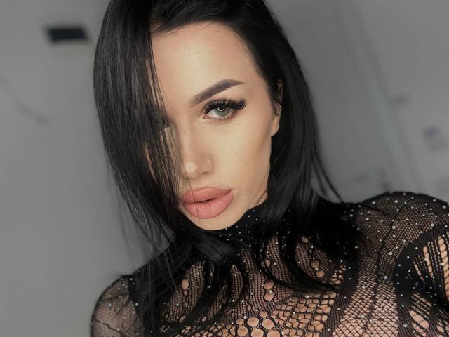 JennaDay with black hair, full lips, and green eyes, wearing a sheer black lace top, looking seductively at the camera, her face partially covered by her hair.
