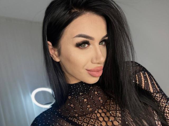 JennaDay with long black hair, green eyes, and full pink lips wearing a black lace top, showing her tits through the patterned fabric. Her makeup is perfect.