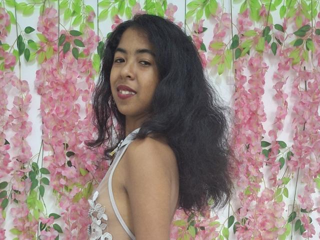 Rosiia with dark curly hair, medium brown skin, wearing a white floral dress, smiling at the camera, pink flower garland in the background. Boobs visible through dress.
