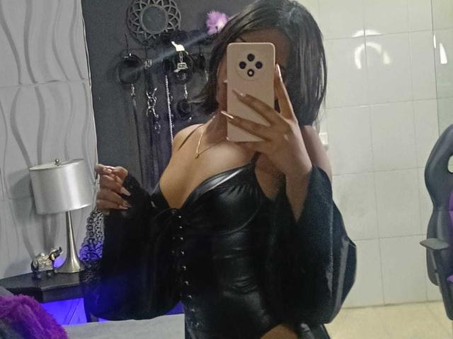 HannaCrow with medium tits, black leather top, black long-sleeve shirt, holding pink phone, covering face, mirror selfie, dark wavy hair, white-tiled wall, black and purple background.
