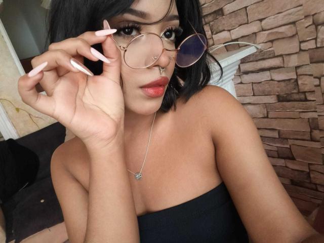 HannaCrow, com cabelo curto preto, piercing dourado no nariz e óculos de sol rosa, segurando copos com unhas longas e rosa, vestindo um top preto sem alças, colar com um pequeno pingente, seios médios..