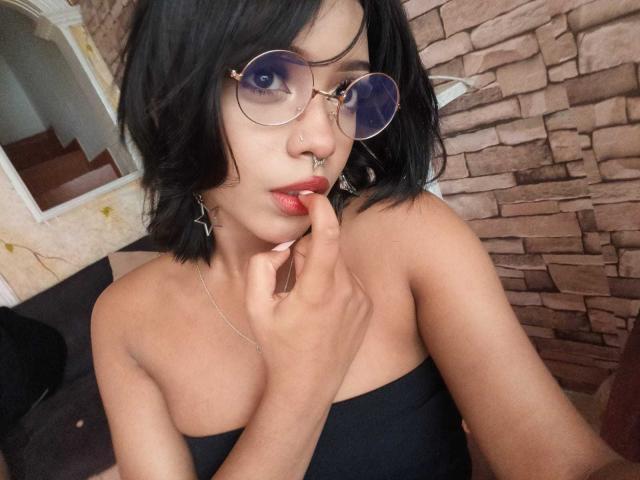 HannaCrow, com cabelo preto, usando óculos redondos, um piercing no nariz e batom vermelho, toca seus lábios vermelhos com o dedo, vestindo uma blusa preta com alças finas, mostrando seus seios..