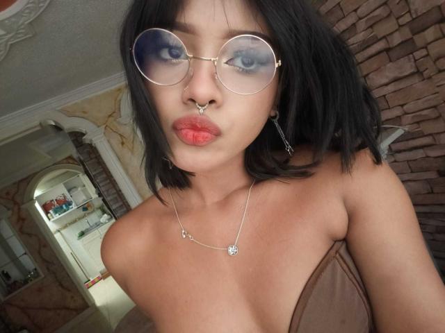 HannaCrow with round glasses, nose ring, and red lipstick, showing off her small tits in a brown strapless top, wearing a necklace with a flower pendant.