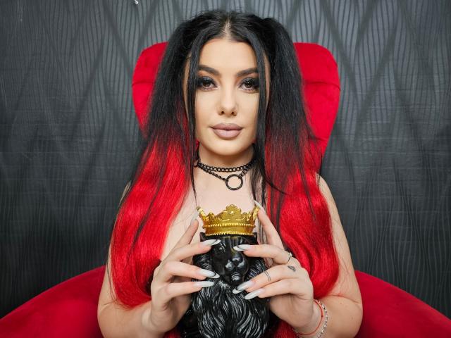 MelissaGlow with red and black hair, holding a golden crown, wearing a black choker, sitting on a red chair, dark backdrop, pale skin, subtle makeup, manicured nails.