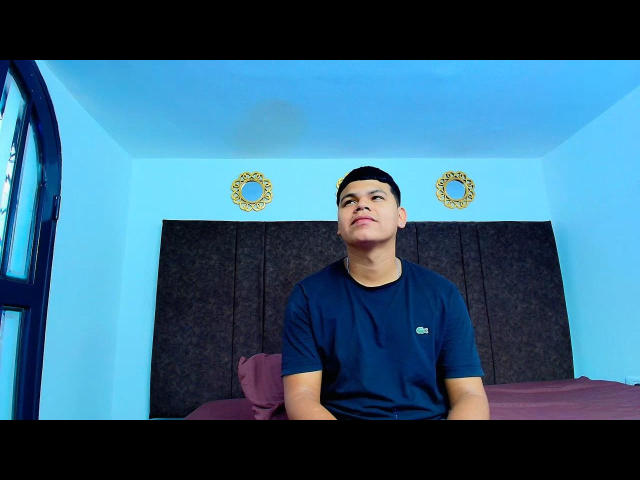 CanelaBoby, short black-haired dude, wearing a dark blue t-shirt, looking up with a slight smirk, standing in front of a dark headboard, two gold circular wall decorations above.