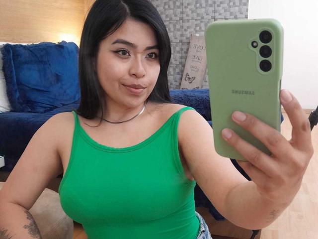 Jesica Rossi tomando un selfie, con una camiseta verde, mostrando sus pechos, sosteniendo un teléfono Samsung verde, expresión de labios fruncidos, cabello negro, suelo de madera, tatuaje en el antebrazo derecho..