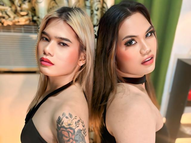TransDuoFullOfLoad with blonde and brown hair, black choker, and tattoos, showing off their tits in a black halter top, posing back-to-back with a sultry look.