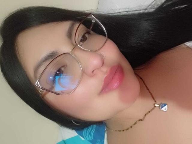 NathDawnson has shiny black hair, glasses, pink lipstick, and a nose ring. She's wearing a necklace with a blue heart pendant. Her skin is light, and she's smiling.