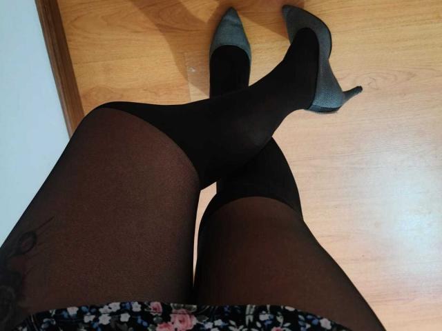NathDawnson's legs in black sheer pantyhose, crossed at the ankles, wearing black and grey high heels, with a floral skirt visible at the bottom. Wooden floor background.