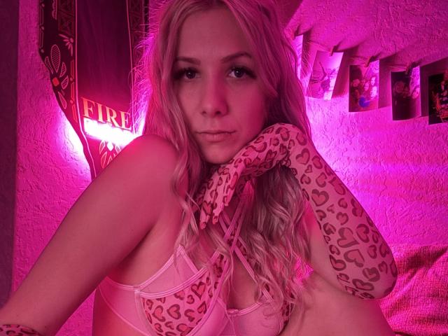 LapinaAnna met roze handschoenen met luipaardprint, een roze beha en lang blond haar, met een frons en haar hand op haar wang, in roze neonlicht, waarbij haar borsten zichtbaar zijn..