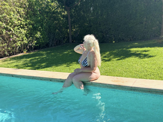 KamaliaSim with big ass, sitting by the pool, wearing a zebra bikini, blonde hair, holding her face, blue water, green grass, and bushy trees in the background.