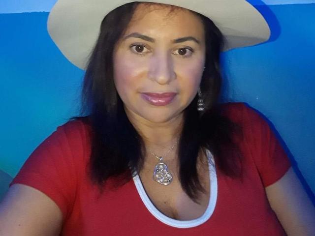 SherahaJade, met bruine ogen, draagt een witte hoed, een rood shirt en een zilveren ketting met een hanger. Ze heeft glanzende, roze lippen en zwart haar van gemiddelde lengte, en ze toont een subtiele glimlach..