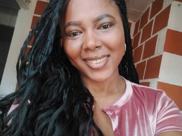 MacarenaBoobs has dark skin, long black braids, and is smiling with glossy pink lips. She's wearing a pink velvet shirt, and has hoop earrings.