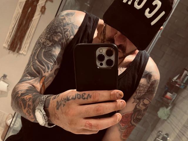 Tätowierte Person, "SexOnMyLeVeL" - sexy, macht ein Selfie, trägt eine schwarze Mütze, ein schwarzes Tanktop und eine silberne Uhr, hält ein Telefon mit "drei" Kameras und zeigt eine tätowierte Hand..
