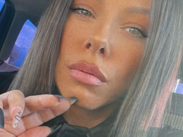 SexOnMyLeVeL-hot heeft donkerbruine huid, lang grijsbruin haar, volle roze lippen en raakt haar onderlip aan met een hand die een kleine zwarte tatoeage heeft..