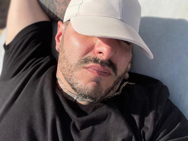 SexOnMyLeVeL - uma pessoa atraente vestindo um boné branco, camisa preta e com tatuagens, com barba e bigode, deitada, com os olhos cobertos pelo boné, iluminada pelo sol, usando um colar de corrente, com a boca ligeiramente aberta..