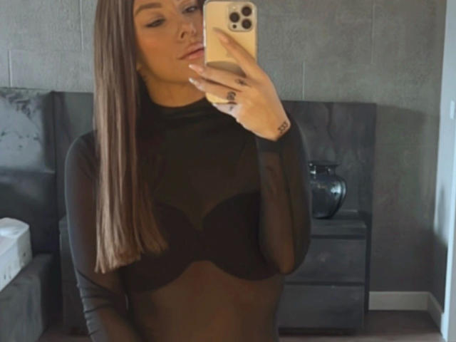 SexyOnMyLeVeL: una ragazza attraente con lunghi capelli castani, che indossa una maglia nera trasparente, che mostra i suoi piccoli seni, tiene un telefono, è in piedi in una stanza grigia e sta guardando lo specchio..