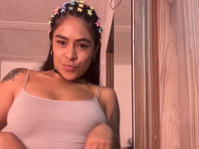 StellaBeckker with a flower crown, wearing a white tank top, showing her small tits and tattooed arm, pouting her lips, in a slightly blurry selfie.