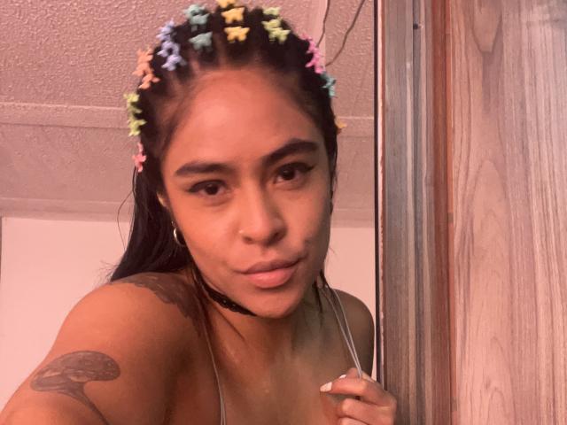 StellaBeckker with colorful braided crown, wearing choker, holding tank top strap, showing tattoo on left shoulder, looking at camera, medium skin, brown eyes, slight pout, wooden door.