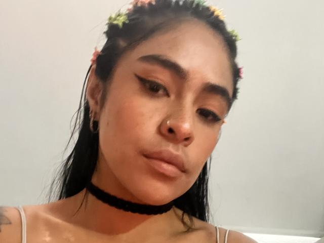 StellaBeckker with dark hair, flower crown, black choker, nose ring, and eyeliner, looking down with pouty lips, wearing a strap dress, visible tattoo on left shoulder.