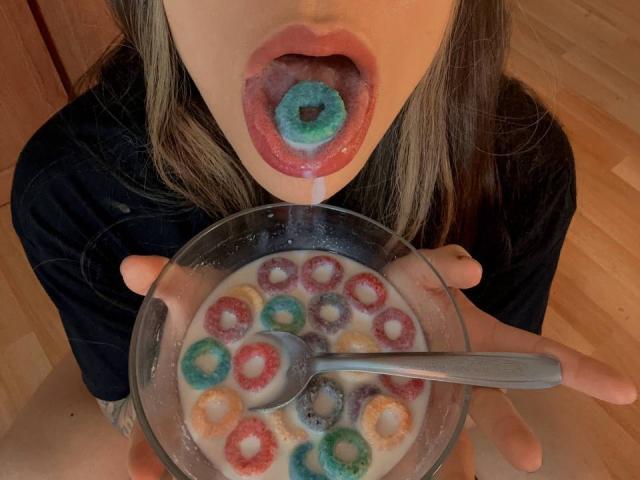 AvaZamora's mouth is open, with a blue donut in her mouth, holding a bowl of colorful donut holes in milk, using a spoon. Her lips are wet.