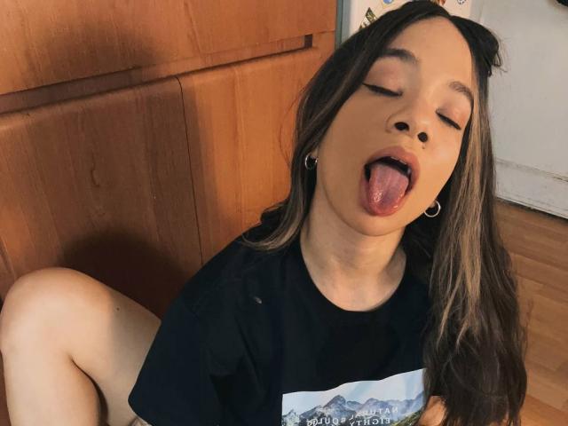 AvaZamora with eyes closed, tongue out, wearing a black shirt, small hoop earrings, and cat ears, sitting on wooden floor, showing her bare thighs and dark brown hair.