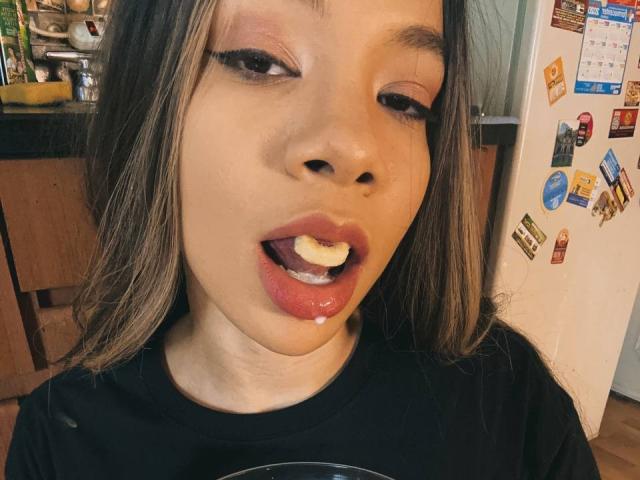 AvaZamora's face with half-licked red lips and a small, white, egg-yolk-like substance on her tongue, wearing black eyeliner, in a kitchen with a fridge in background.