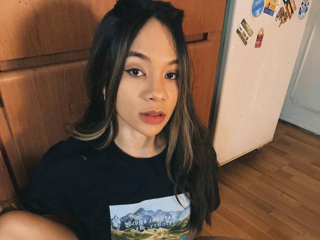 AvaZamora with long brown hair, black shirt, and mountain graphic, sitting on wooden floor, looking at camera with slightly parted lips, fridge with stickers in background.