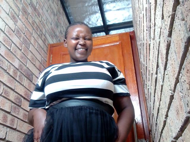 ShyMuse with dark skin, smiling, wearing a black and white striped shirt, black skirt, and belt, standing in a brick hallway, showing her big ass.