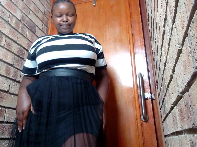 ShyMuse with dark skin, wearing a black and white striped top, black skirt, and belt, smiling, standing in front of a wooden door, hands on her skirt.