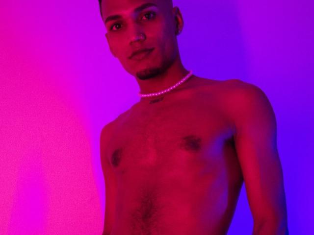 AlejoMorgan with a dark skin tone, short hair, and a necklace, shirtless, showing a muscular chest and small, dark nipples, illuminated by pink and purple light.