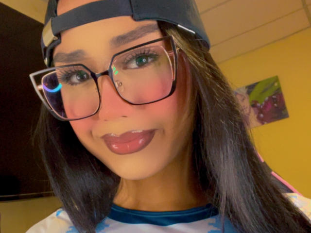 AnielFetish has dark hair, glasses, a black cap, and glossy pink lips. Her face is slightly flushed, with a cute nose piercing. She's wearing a colorful shirt.