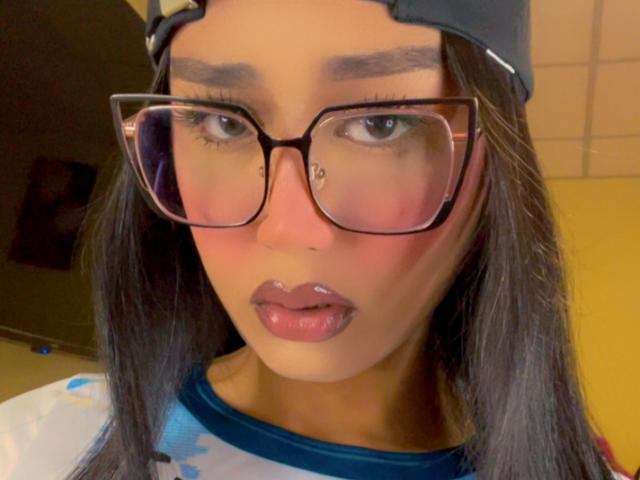 AnielFetish has dark brown eyes, thick eyebrows, and glossy pink lips, wearing black-rimmed glasses and a blue-and-white shirt, with a black cap tilted back.