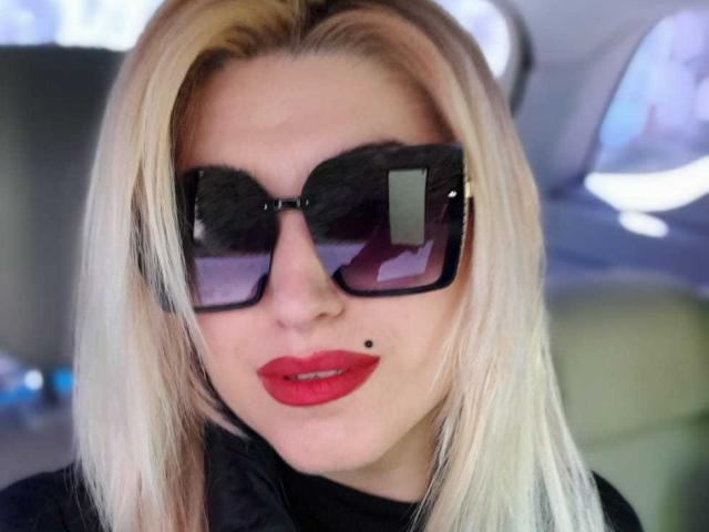 ElizabethRaegan with blonde hair, black sunglasses, red lipstick, and a small mole under her right eye, wearing a black top, looking directly at the camera.