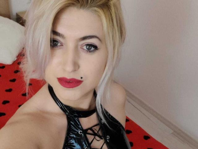 Elizabeth Raegan mit blonden Haaren, rotem Lippenstift, einem schwarzen Vinyl-Haltertop und einem Muttermal auf der Oberlippe, die ihre Brüste und ihr hübsches Gesicht in einem Selfie zur Schau stellt..