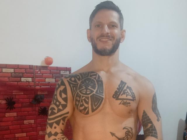 Ein tätowierter Samurai ohne Hemd, mit muskulöser Brust, kurzen, dunklen Haaren und Bart, lächelnd. Im Hintergrund eine rote Backsteinmauer und schwarze Spinnendekorationen. Seine Tattoos umfassen ein großes Motiv auf der Brust sowie Designs auf den Armen..