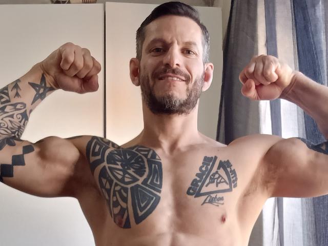 Samuraji é um sujeito musculoso, tatuado e com barba, que exibe seus bíceps e sorri. Ele tem uma tatuagem triangular no peito e uma tatuagem geométrica no peito..