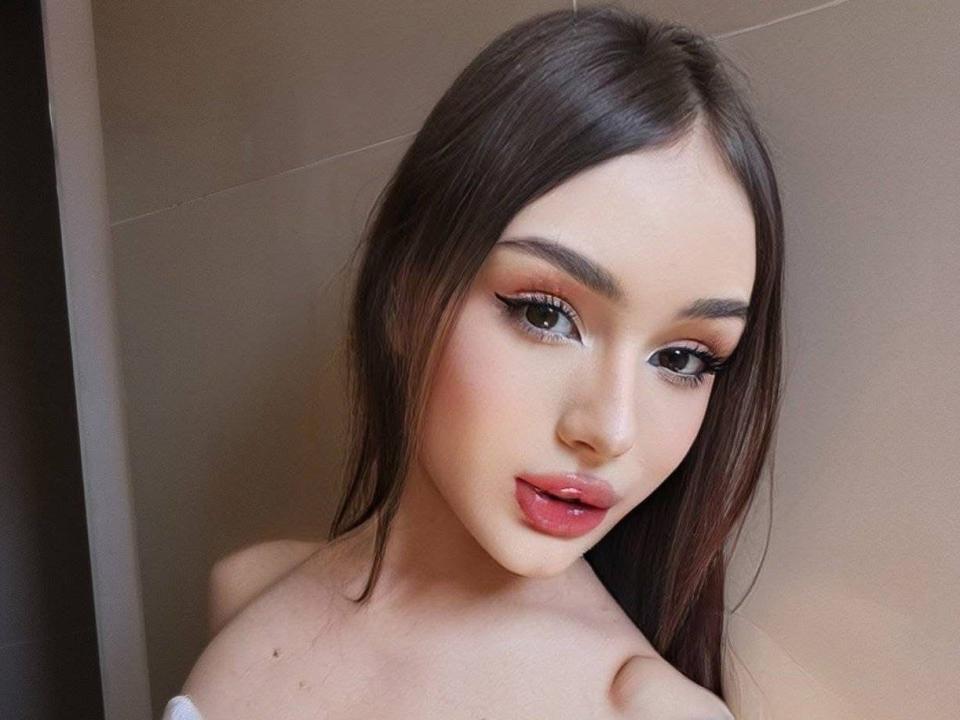 DecameronLuxury has long brown hair, green eyes, and red lipstick. She's looking at the camera with a slight smile, showing off her cute, sexy face and small tits.
