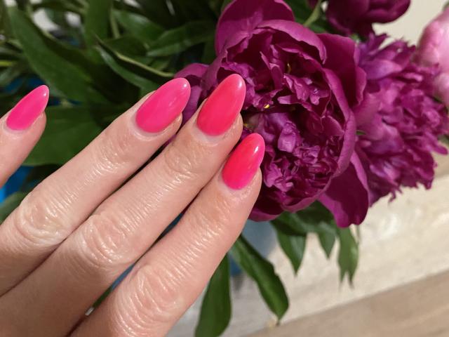 AlicePrings' Hand mit glänzenden, rosafarbenen Nägeln, die lila Pfingstrosen hält. Ihre Finger sind leicht runzelig, und im Hintergrund sind grüne Blätter zu sehen..