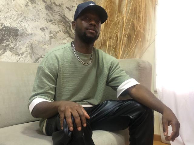 JacksonBlaze, dark-skinned dude with a beard, wearing a green sweater, black leather pants, and a navy cap, sitting on a gray couch, looking slightly to the side.