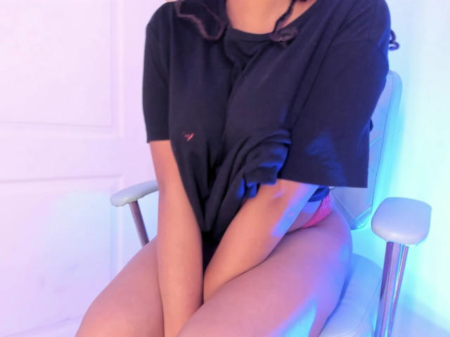 JadeFoxmei sitting with her black shirt covering her tits, pink panties showing her ass, and her legs together on a white chair, in soft blue and pink lighting.