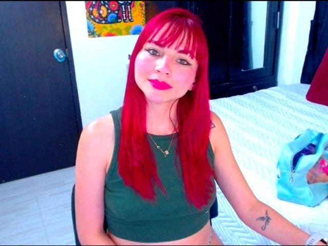 EmelySofia with bright red hair, green crop top, and red lipstick, sitting on a bed with white sheets, showing a small tattoo on her right forearm.