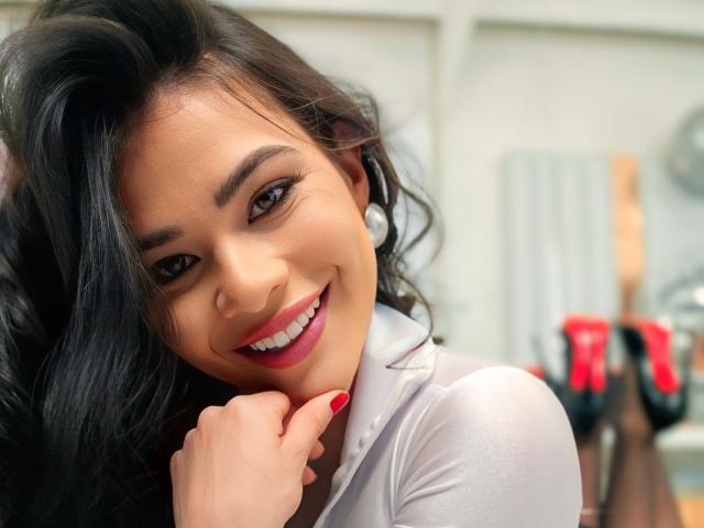 PaulinaValley has dark wavy hair, brown eyes, and a cute smile. She's wearing a white shirt with pearl earrings, and has red nail polish. Her hand is near her face.