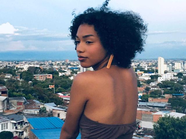 Ayla Calloway, avec ses cheveux noirs bouclés, sa peau brune et ses boucles d'oreilles dorées, montrant son dos nu et son haut marron sans bretelles, regardant par-dessus son épaule, avec un paysage urbain en arrière-plan..