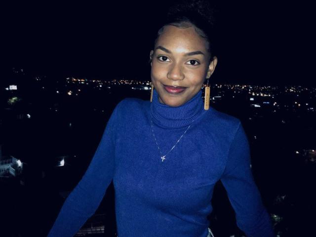 AylaCallowey with dark skin, smiling, wearing a blue turtleneck, gold earrings, and a silver necklace, standing at night with city lights in the background.