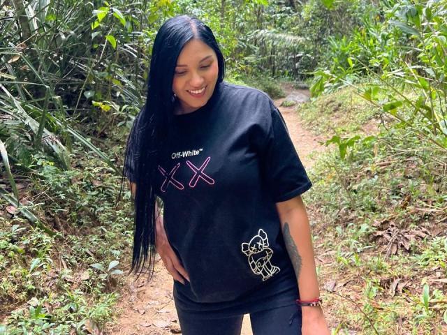 YessiJaymes with long black hair, smiling, wearing a black tee with pink "X"s and a small white drawing, standing on a dirt path in a green forest.