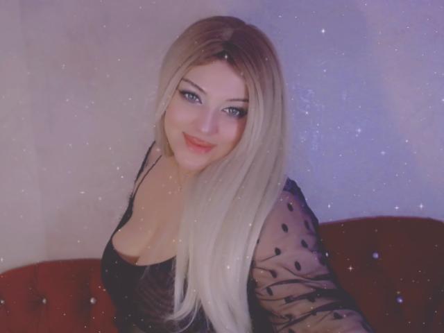 SecretLand hat blondes Haar, blaue Augen und ein süßes Lächeln. Sie trägt ein schwarzes, durchsichtiges Oberteil, das ihre Brüste zeigt, und sitzt auf einem roten Sofa. Ihr Make-up ist verführerisch und sie ist sehr attraktiv..
