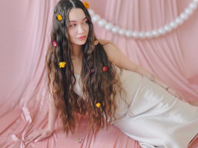 LilianCat-hot with long wavy brown hair, yellow and pink flower clips, wearing a shiny silver dress, leaning on pink fabric, smiling softly. Boobs partially visible under dress.