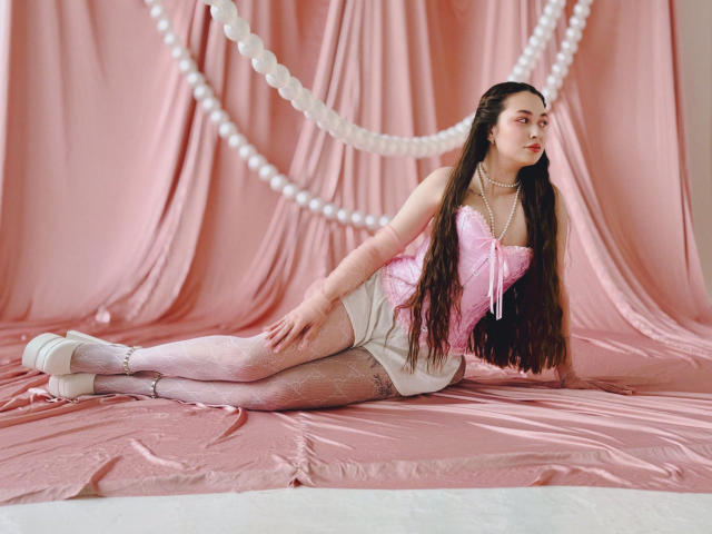 LilianCat-hot with long brown hair, pink corset, white lace stockings, and pearl necklace, lying on pink satin, wearing white heels, looking to the side, with white pearls in background.